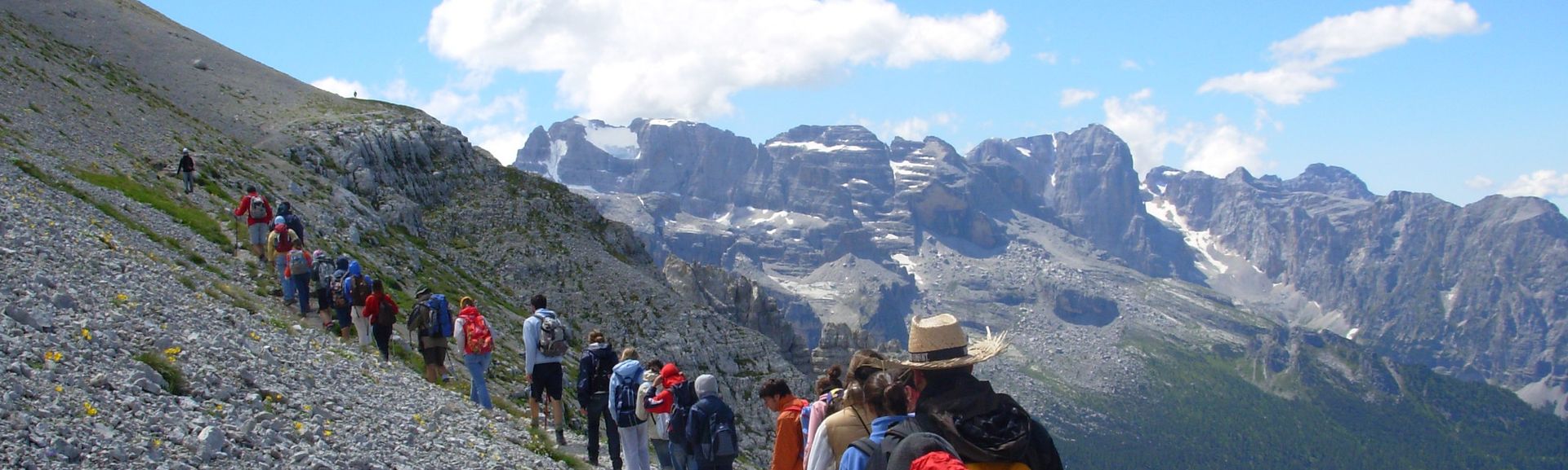 Una fila di giovani della Comunità cammina su un sentiero di montagna davanti alle Dolomiti di Brenta